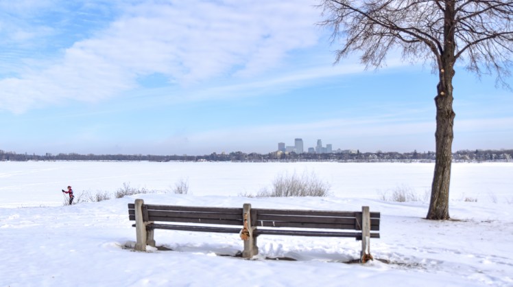 Park bench in snow by frozen lake with skier and skyline